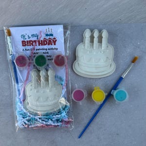 May include: A DIY birthday-themed painting kit. The kit includes a white cake-shaped ceramic piece with three candle-shaped details, paint pots in pink, yellow, and blue, and two paintbrushes. The packaging reads "It's my BIRTHDAY".