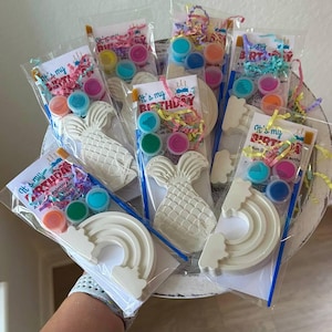 May include: Several clear plastic bags containing paint kits with the text "It's my BIRTHDAY". Each kit includes a paintable mermaid, rainbow, or cake shape, paint pots, and a paintbrush. The kits are arranged in a decorative basket.