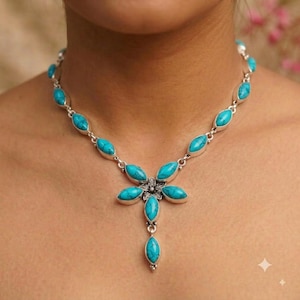 Puede incluir: Un collar de tono plateado que presenta una serie de piedras turquesa en forma de marquesa unidas entre sí. El centro del collar muestra una disposición en forma de flor de cinco piedras turquesa con un pequeño detalle floral plateado.