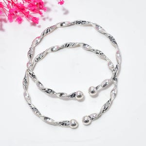 May include: Two silver-toned metal bangles featuring a twisted, rope-like design with intricate engraved patterns along the surface. Each bangle is an open-ended cuff style, finished with a smooth, polished spherical bead at both ends of the band.