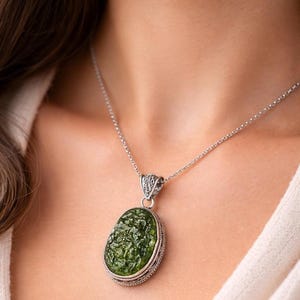 May include: A silver pendant necklace featuring an oval-shaped, textured dark green stone set in a decorative silver frame with intricate metalwork on the bail, suspended from a delicate silver chain that rests against the collarbone area.