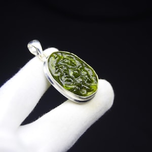 May include: A green pendant with a silver-colored bezel. The pendant has an oval shape and a textured surface. The background is black.