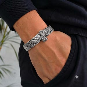 Puede incluir: Un brazalete de metal en tono plateado con un diseño detallado de un águila en relieve con las alas extendidas. El brazalete tiene un acabado texturizado y oxidado que resalta los intrincados patrones de plumas en toda la superficie.
