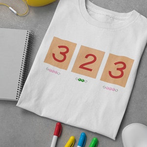 May include: White t-shirt with the numbers 323 printed on it in red. The numbers are on brown squares with pink and green beads underneath.