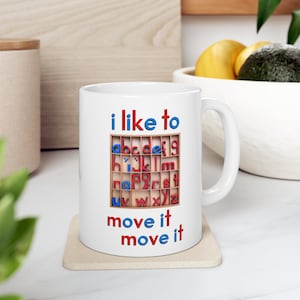 May include: White ceramic mug with a red and blue alphabet block design. The mug reads "i like to move it move it".