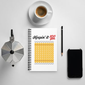 May include: A white spiral-bound notebook with the text "Keepin' it 100" and a red emoji. The notebook features a grid of yellow circles. A cup of coffee, a pencil, a phone, and a coffee maker are also on the white surface.
