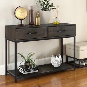 May include: A dark brown console table with two drawers and a lower shelf, supported by a black metal frame. The table is decorated with a globe, bottles, plants, books, and a decorative vase. The table is 122 cm long.