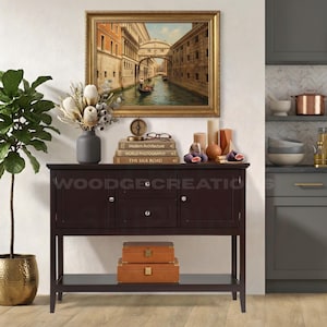 May include: A dark brown console table with a framed painting of a canal scene. The table is decorated with a vase of dried flowers, books, candles, and figs. Two leather boxes are on the bottom shelf. The text "WOODGE CREATIONS" is visible.