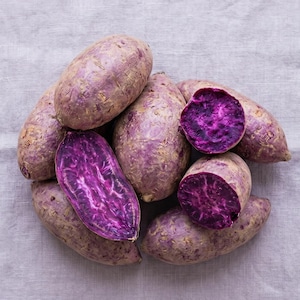 Könnte beinhalten: Eine Ansammlung von lila Süßkartoffeln, einige ganz und andere aufgeschnitten, um das leuchtend violette Fruchtfleisch zu zeigen. Die Kartoffeln haben eine hellviolette Schale mit leicht rauer Textur. Die Schnittflächen zeigen eine tiefe, satte violette Farbe.