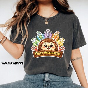 Può includere: T-shirt grigio scuro con un disegno di un riccio dei cartoni animati circondato da siringhe colorate. Il riccio ha un'espressione amichevole e il testo "Fully Vaccinated" è su un cartello di legno. Uno stile casual e confortevole.
