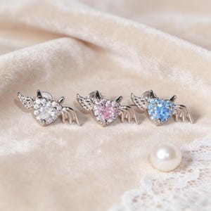 以下が含まれることがあります： 羽と角が付いた、3つの銀色のハート型イヤリング。それぞれに異なる色の中心石があります。クリア、ピンク、ブルー。白い真珠がクリーム色の表面の横にあります。イヤリングはおそらくピアス用です。