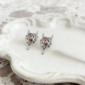 以下が含まれることがあります： シルバーカラーのスタッドピアス。各ピアスは、中央に赤い宝石があり、曲がった角のある詳細なデザインで囲まれています。ピアスには、中央のデザインの下にユニークなドロップのような要素があります。