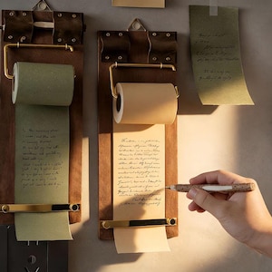 Könnte beinhalten: Zwei Holz-Notiztafeln mit Papierrollen, Messingbeschlägen und handschriftlichen Notizen. Eine Tafel hat eine grüne Papierrolle, die andere eine beige Rolle. Eine Hand hält einen Stift, bereit, auf das Papier zu schreiben.