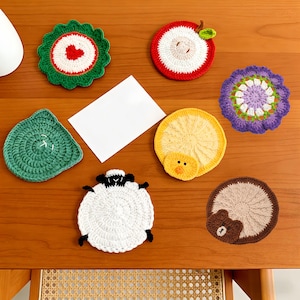 May include: A collection of handmade crochet coasters in various shapes and colors, including a green leaf, a red apple, a purple flower, a yellow chick, a white sheep, a brown bear, and a green coaster with a heart.