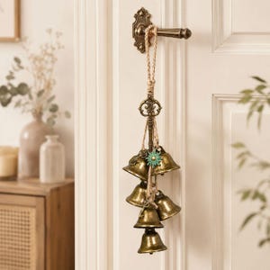 May include: A decorative hanging ornament featuring a vintage-style brass key and five brass bells suspended by a braided jute rope. A small turquoise-colored sun charm is attached to the rope, and the entire piece hangs from a brass door handle.