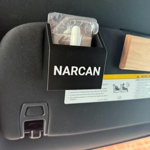 May include: A black rectangular holder attached to a vehicle sun visor, featuring the white text NARCAN printed on the front. The holder contains a clear plastic package with a white nasal spray device visible inside the compartment.