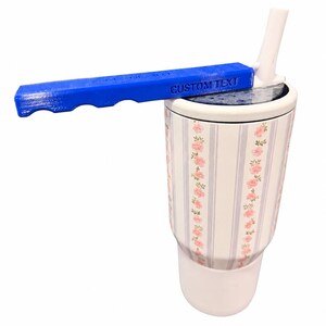 May include: A white tumbler with a floral pattern of pink roses and light blue vertical stripes, featuring a white lid with a straw and a blue handle attachment that has the words CUSTOM TEXT embossed on the side of the handle surface.