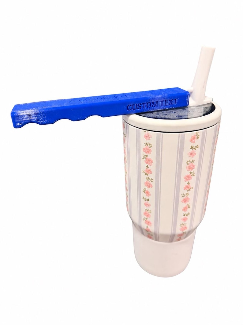 May include: A white tumbler with a floral pattern of pink roses and light blue vertical stripes, featuring a white lid with a straw and a blue handle attachment that has the words CUSTOM TEXT embossed on the side of the handle surface.