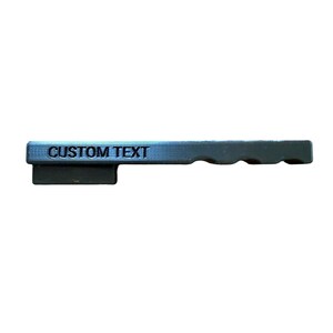 May include: A matte black, rectangular, 3D-printed object featuring the words CUSTOM TEXT embossed on the side. The item has a flat, elongated top section with a smaller, rectangular base attached to the left side and a series of three rounded indentations along the right edge.