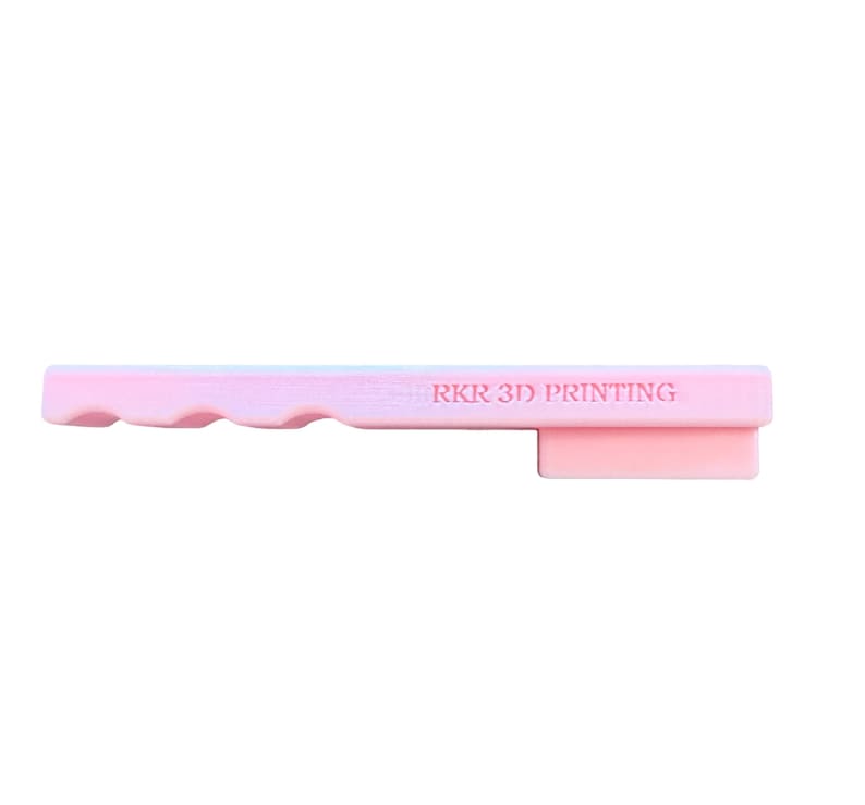 May include: A light pink, elongated, rectangular plastic object with a wavy edge on one side and a protruding rectangular tab on the other. The text RKR 3D PRINTING is embossed in a horizontal line across the center of the flat surface.