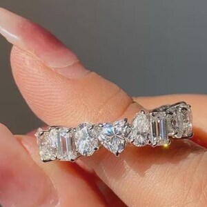 Könnte beinhalten: Ein silberner Ring mit einer Reihe klarer, funkelnder Edelsteine in verschiedenen Formen, darunter Smaragd-, Oval-, Tropfen- und herzförmige Steine. Der Ring wird von einer Person mit hellem Hautton gehalten.