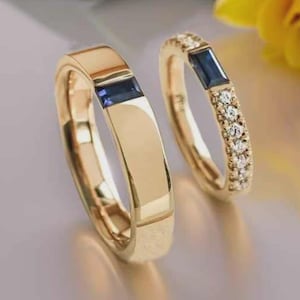 Può includere: Due fedi nuziali d'oro. Un anello presenta una gemma blu rettangolare, mentre l'altro ha una gemma blu simile ed è incrostato di piccole gemme chiare e rotonde. Gli anelli sono su uno sfondo morbido e sfocato.