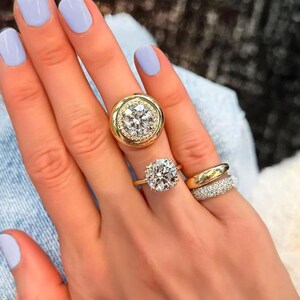 Op de afbeelding: Een hand toont verschillende gouden ringen met heldere edelstenen. Eén ring heeft een grote, ronde diamant omringd door kleinere stenen. Een andere ring heeft een enkele, prominente diamant. Een derde ring is een eenvoudige gouden band.