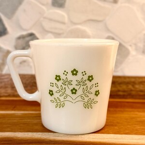May include: White ceramic mug with a green floral design. The mug has a handle and a slightly flared shape. The floral pattern includes leaves and small flowers. The mug is set on a wooden surface.