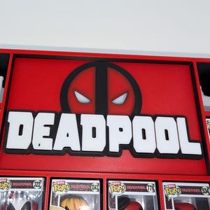 Può includere: Uno scaffale espositivo rosso e nero con il logo Deadpool in lettere bianche e nere. Il pannello centrale mostra l'iconico emblema della maschera rossa e nera. Sotto e ai lati ci sono scomparti rettangolari contenenti varie statuette Deadpool.