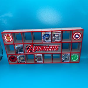 Peut inclure: Une étagère de présentation rectangulaire rouge et grise avec le logo Avengers au centre. L'unité comporte plusieurs compartiments carrés, certains contenant de petites figurines de collection en boîte, tandis que d'autres arborent des icônes circulaires.