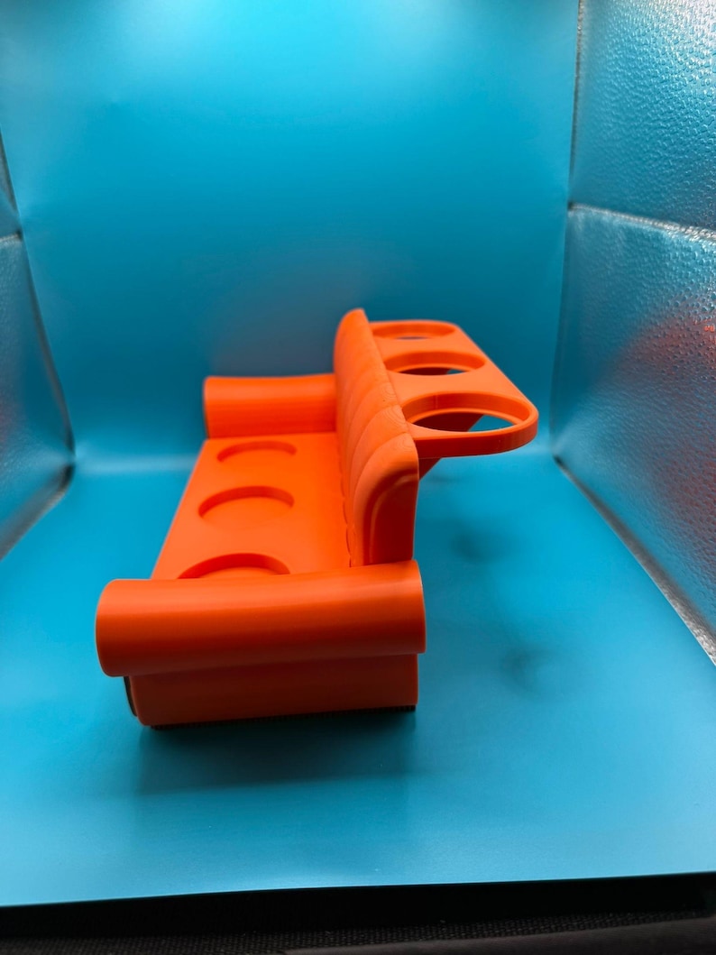 May include: Bright orange plastic cup holder with four circular openings. The holder has a curved design with a back and a base, and is designed to hold cups or bottles. The holder is on a blue background.