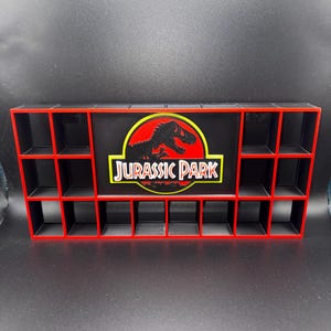 Peut inclure: Une étagère de présentation rectangulaire noire et rouge dotée de multiples petits compartiments carrés. Le panneau central affiche le logo Jurassic Park, comprenant une silhouette noire de T-Rex dans un cercle jaune avec le texte Jurassic Park.