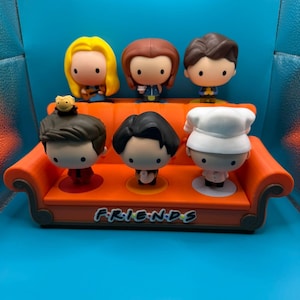 May include: A set of six Friends character figurines displayed on an orange couch-shaped stand. The figurines have large heads and small bodies, with distinct hairstyles and outfits. The stand has the word "FRIENDS" in white lettering.