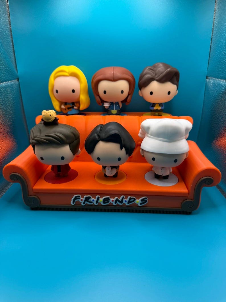 May include: A set of six Friends character figurines displayed on an orange couch-shaped stand. The figurines have large heads and small bodies, with distinct hairstyles and outfits. The stand has the word "FRIENDS" in white lettering.
