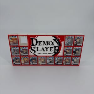 Peut inclure: Une étagère de présentation rectangulaire blanche et rouge avec le logo Demon Slayer Kimetsu no Yaiba au centre. L'unité comporte plusieurs compartiments carrés individuels, chacun contenant une petite figurine dans son emballage d'origine.