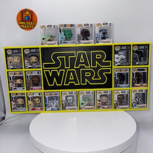 May include: A yellow rectangular display shelf featuring a black centre panel with the yellow Star Wars logo. The shelf holds fourteen Star Wars Funko Pop figures in their original boxes, with four additional figures displayed on top of the unit.