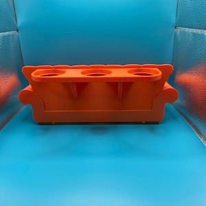 May include: An orange, miniature sofa-shaped drink holder with three circular cup holders. The design includes armrests and a base, resembling a small couch. The item is made of a solid, opaque material.