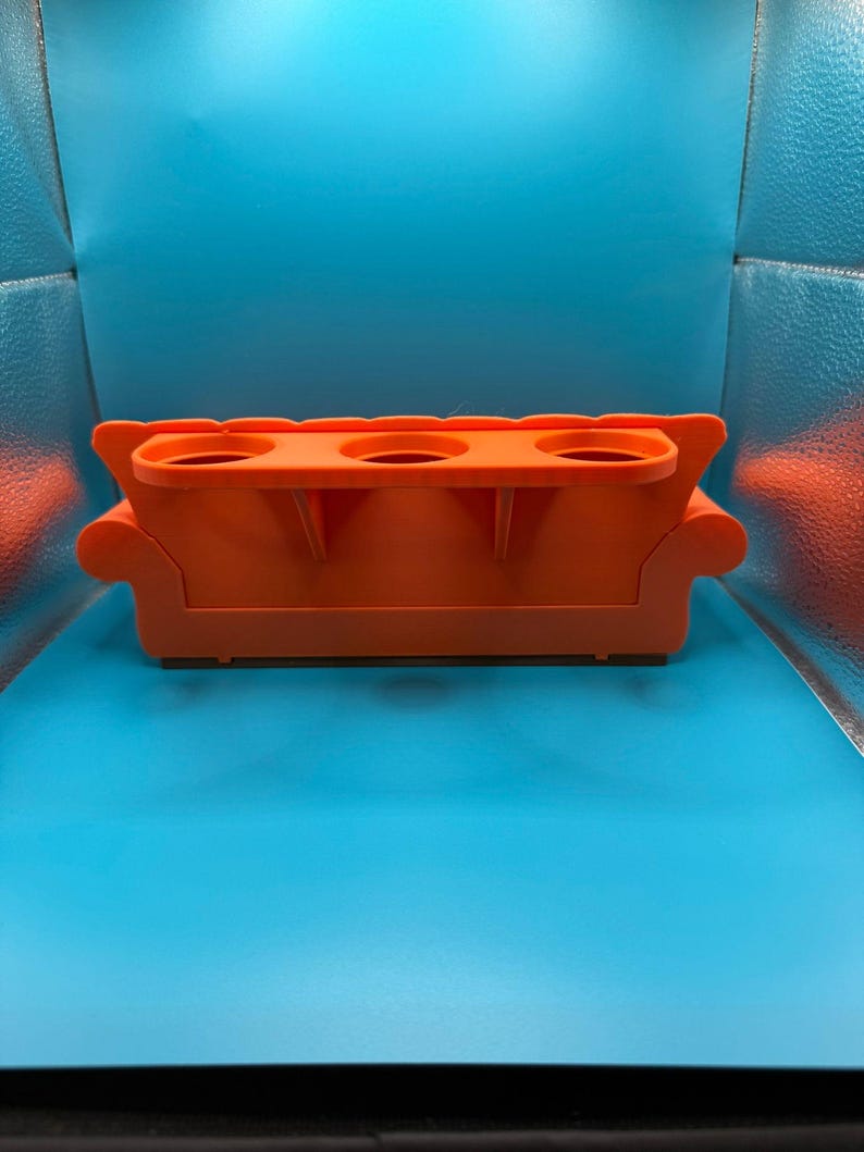 May include: An orange, miniature sofa-shaped drink holder with three circular cup holders. The design includes armrests and a base, resembling a small couch. The item is made of a solid, opaque material.