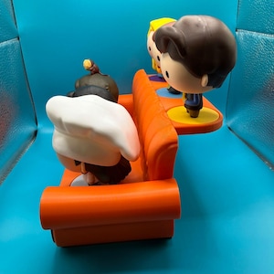 May include: A Funko Pop! vinyl figure set featuring characters from the TV show Friends. The set includes figures of the characters sitting on an orange couch and standing on a yellow platform. The figures have large heads and stylized features.