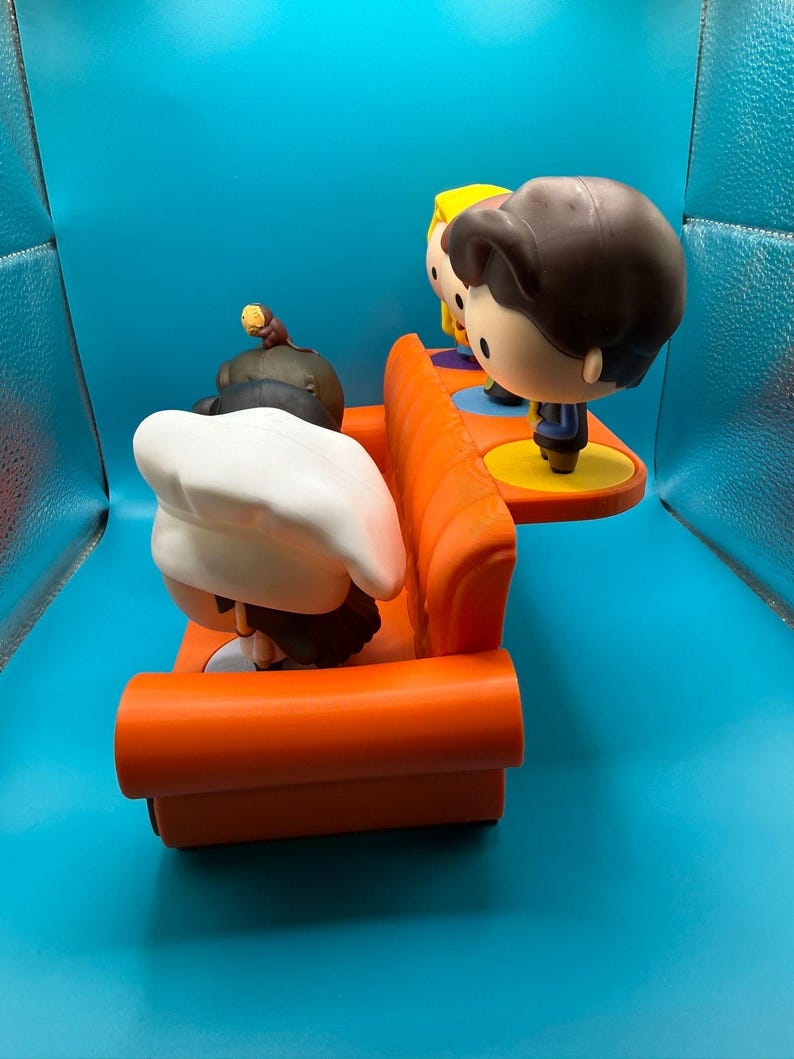 May include: A Funko Pop! vinyl figure set featuring characters from the TV show Friends. The set includes figures of the characters sitting on an orange couch and standing on a yellow platform. The figures have large heads and stylized features.