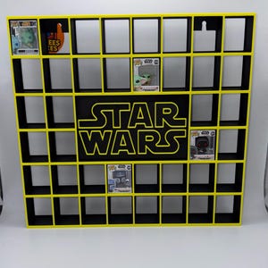 Peut inclure: Une étagère de présentation rectangulaire noire avec un cadre jaune vif comportant une grille de compartiments carrés. Le panneau central affiche le logo Star Wars en texte jaune. Quatre figurines de collection Star Wars sont placées.