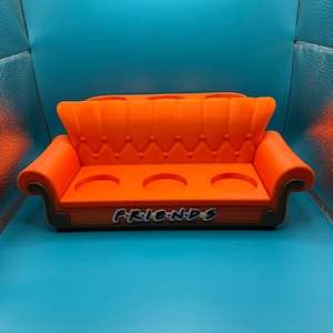 May include: An orange, miniature couch-shaped drink holder with three circular cup holders. The word "FRIENDS" is printed on the front in the show's iconic font. The couch has a tufted back and curved armrests, and is set against a blue background.