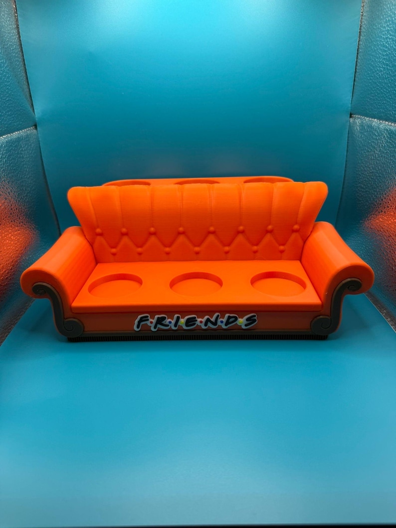 May include: An orange, miniature couch-shaped drink holder with three circular cup holders. The word "FRIENDS" is printed on the front in the show's iconic font. The couch has a tufted back and curved armrests, and is set against a blue background.