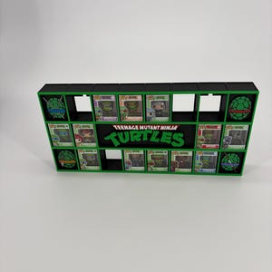 May include: A black and green display shelf featuring the Teenage Mutant Ninja Turtles logo in the centre. The unit has multiple rectangular compartments, some containing small collectible figures in boxes and others displaying turtle shell icons.