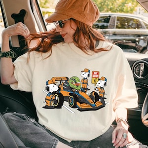 May include: Cream-colored t-shirt featuring a cartoon illustration of Snoopy and a Formula 1 race car. The car is orange and blue, with Snoopy characters in racing attire. The text "PIT STOP LAP 12" is on a sign.