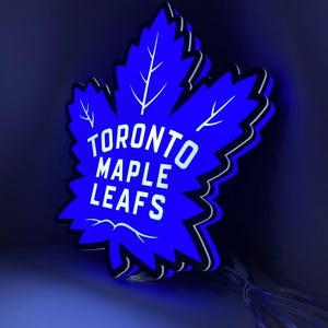 Puede incluir: Un letrero azul en forma de hoja de arce con las palabras "TORONTO MAPLE LEAFS" en blanco. El letrero tiene un contorno negro y vetas blancas, y está iluminado con un brillo azul. Un cable se extiende desde la parte inferior.