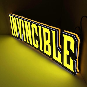 Könnte beinhalten: Leuchtschild mit dem Wort "INVINCIBLE" in fetten, gelben Buchstaben. Die Buchstaben haben eine schwarze Umrandung mit einem weißen Rand, was einen starken Kontrast erzeugt. Das rechteckige Schild strahlt hell, was auf eine interne Beleuchtung hindeutet.