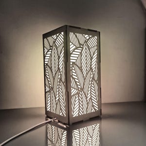Peut inclure: Une lampe rectangulaire en bois avec un motif de feuilles découpé sur les côtés. La lampe est éclairée de l'intérieur, projetant une lueur chaleureuse. La lampe est reliée à un cordon d'alimentation blanc.