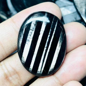 May include: An oval-shaped black gemstone cabochon featuring distinct, parallel silver-white stripes running vertically across its polished, reflective surface. The stone displays a smooth, rounded texture with a high-gloss finish and sharp contrast.