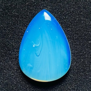 Peut inclure: Un cabochon en forme de goutte d'eau avec une surface lisse et polie. La pierre présente un dégradé bleu translucide qui passe d'un azur profond et saturé sur les bords à un centre blanc laiteux plus clair avec de subtils tourbillons.